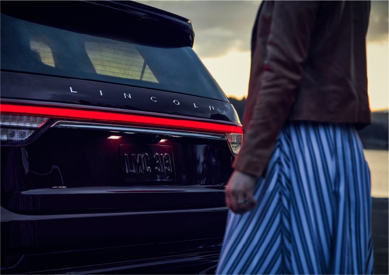 A person is shown near the rear of a 2023 Lincoln Aviator® SUV as the Lincoln Embrace illuminates the rear lights | Ted Russell Lincoln in Knoxville TN